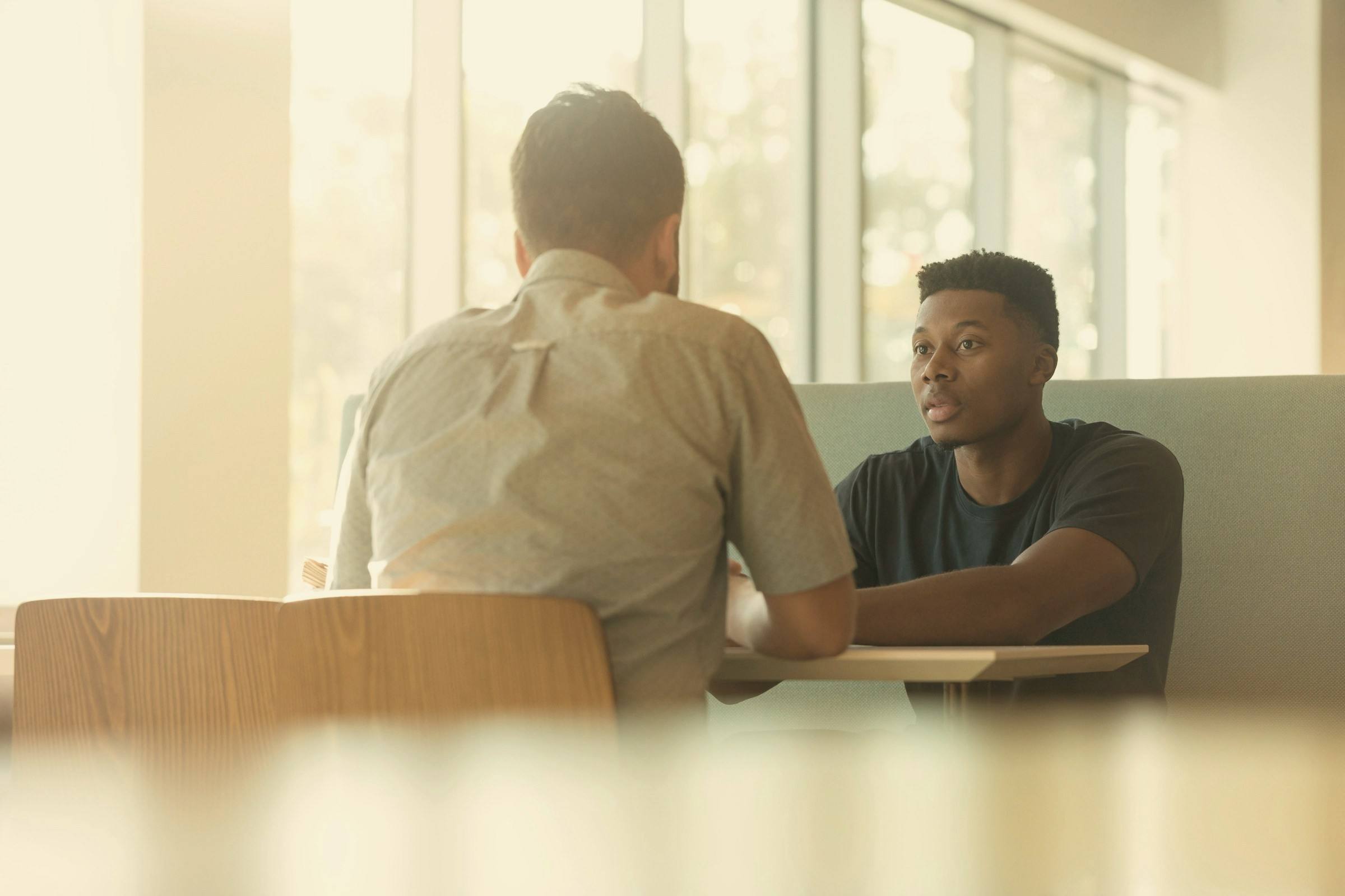 Two men in a focused conversation at a table beside sunlit windows.