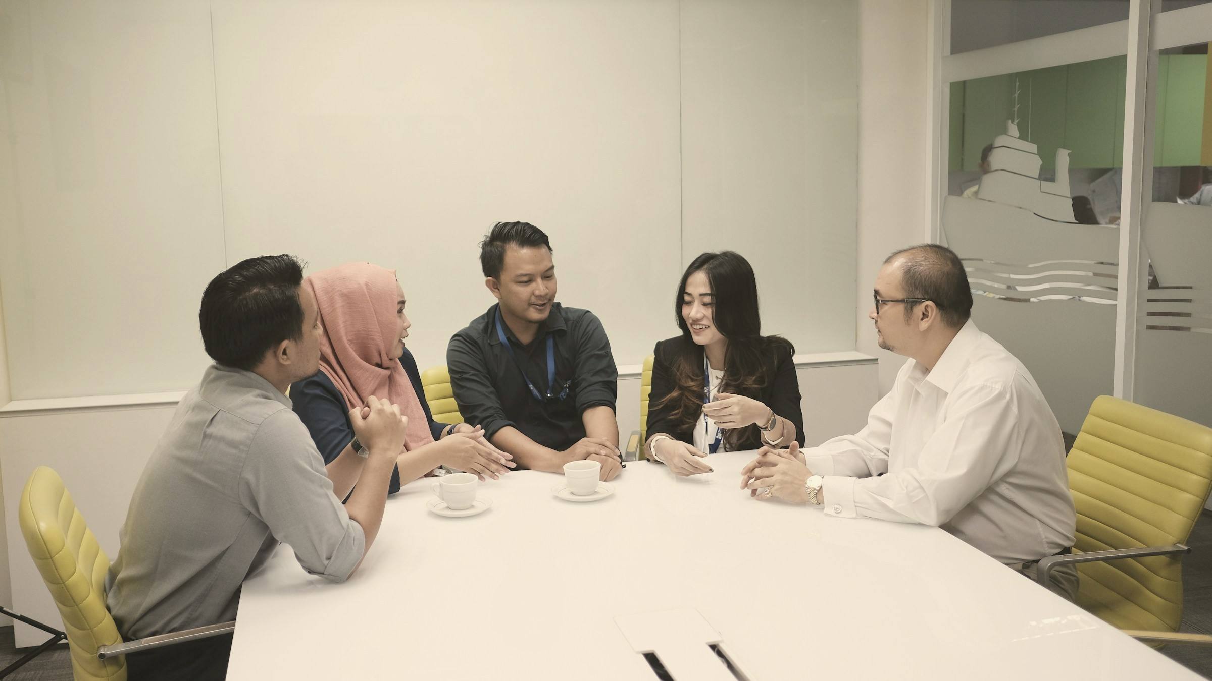Five professionals in conversation around a white meeting table in a bright office.